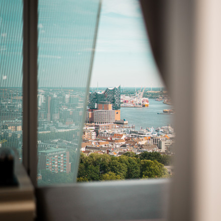 Stadtlandschaft mit Blick auf einen Hafen und ein modernes Gebäude, eingerahmt von einem Fenster und einem beigen Vorhang im Vordergrund.